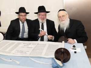 Three men in traditional Jewish attire seated at a table with a Torah scroll, involved in what appears to be a Torah dedication ceremony.