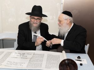 Two men in religious Jewish garb smiling at each other, sharing a quill over an open Torah scroll, signifying participation in a sacred Jewish tradition.
