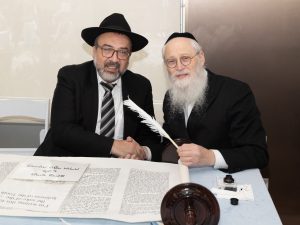 A man in a black hat and a man in traditional Jewish attire holding a quill over a Torah scroll, indicative of a Jewish religious ceremony.
