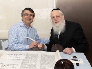 A casual portrait of two men at a table with a Torah scroll, one in a buttoned-up shirt and the other in religious Jewish attire, suggesting a community event.
