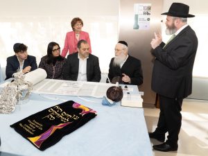 A group seated at a table with a Torah scroll, with an older woman standing behind, and a man speaking, suggesting an educational or religious ceremony.