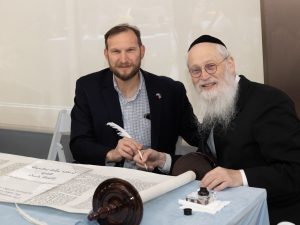 Two men at a table with a Torah scroll, one holding a quill, suggesting a ritualistic act of writing or dedication within a Jewish tradition.