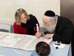 A woman and a man at a table, focused on a Torah scroll with a quill in hand, portraying an engagement in a traditional Jewish ceremonial activity.
