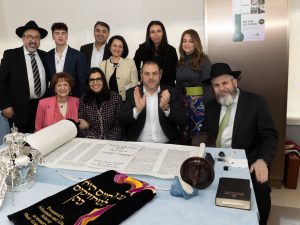 A vibrant group portrait with individuals of various ages around a table with a Torah scroll, indicating a family or community celebration.