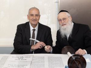 Image of two men sitting at a table with a Hebrew scroll, smiling and poised as if in the middle of a significant cultural or religious signing ceremony.