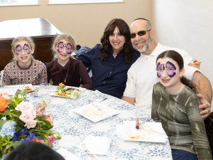 Two girls with butterfly face paint and a couple, all seated at a dining table, sharing a family-like moment at a communal or festive event.