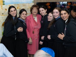 A woman in a pink blazer surrounded by young girls in uniform, smiling for a group photograph in a friendly and joyous environment.