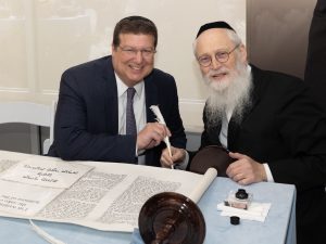 Image of two men sitting at a table with a Hebrew scroll, smiling and poised as if in the middle of a significant cultural or religious signing ceremony.