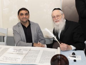 A man in a grey blazer and black shirt sits beside an elderly man with a long white beard wearing a black hat, both seated at a table with a Torah scroll and a quill, suggesting participation in a religious or cultural ceremony.