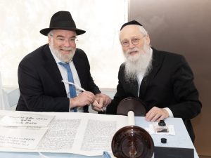 Two smiling men, one in a black hat, sit at a table with a Torah scroll laid out in front of them, one holding a quill, indicating a moment of celebration or honor in a Jewish tradition.