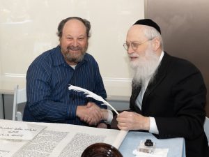 A candid shot of two men at a table, one of them with a quill in hand, engaging in a traditional activity involving a Hebrew scroll, indicative of a religious or cultural ritual.
