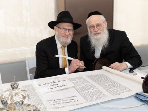 A photograph of two men at a table with a quill and an open Hebrew scroll, displaying elements typical of a cultural or religious ceremonial event.