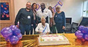 A group of family and friends gather to celebrate Bertie Stanley's 100th birthday at the Margaret Tietz Nursing and Rehabilitation Center. Bertie, wearing a sash and tiara, is seated at the center with a large birthday cake in front of her, surrounded by joyful loved ones and colorful balloons.