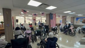 A room decorated with American flags and patriotic bunting, featuring seniors in wheelchairs gathered around a speaker delivering a presentation at a celebratory event