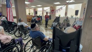 A wide view of a patriotic-themed senior living event, showcasing residents in wheelchairs attentively listening to a speaker under festive American flag decorations.