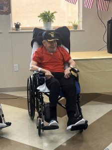 A proud Navy veteran sits in a wheelchair, wearing his service cap during a senior living community event honoring veterans with warm recognition and camaraderie