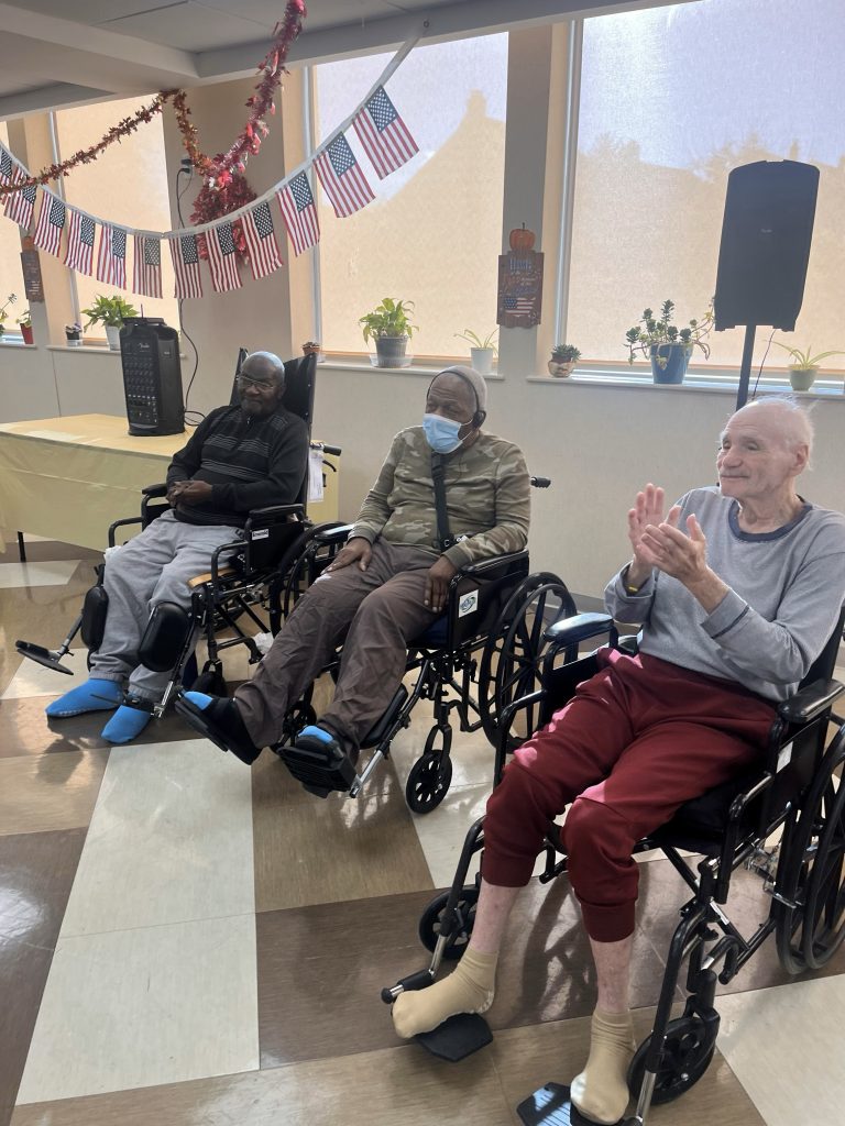 Three senior residents in wheelchairs enjoy a patriotic-themed event, one clapping enthusiastically, as they share in a community celebration honoring veterans and fostering togetherness