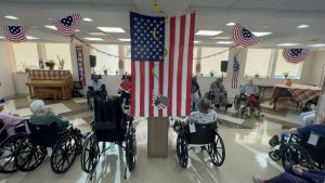 A patriotic-themed event in a senior living facility, with participants seated in wheelchairs facing a large American flag and a decorated podium.