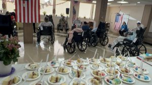 A table filled with plates of colorful cake slices in the foreground, while a group of seniors in wheelchairs enjoy a patriotic-themed gathering adorned with American flags and festive decorations