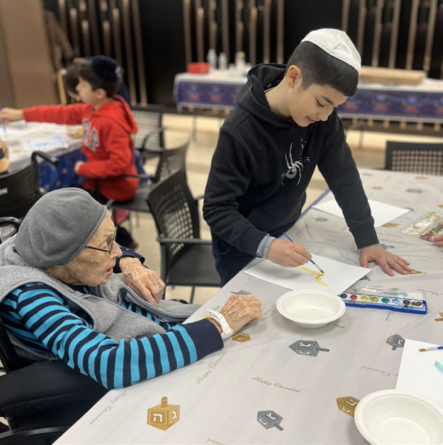 An elderly woman and a young boy sit together at a table covered with a dreidel-themed tablecloth. The boy, wearing a kippah and black hoodie, is painting on a piece of paper with watercolors, while the elderly woman, wearing a gray hat and glasses, watches attentively. Other children can be seen in the background participating in similar activities.